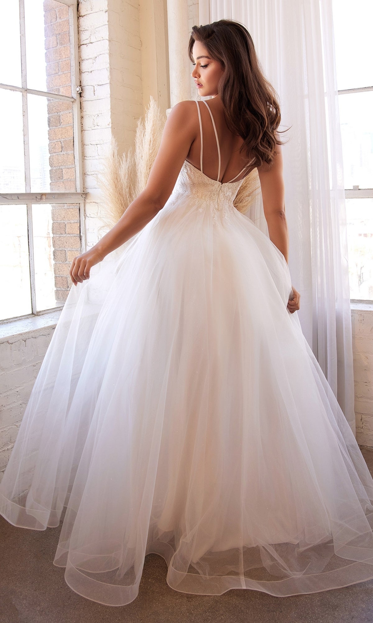 A woman stands indoors by large windows in the Off-White Long A-Line Wedding Dress CD0154W, featuring thin straps, an open back, and a flowing tulle design for an elegant, timeless bridal look.